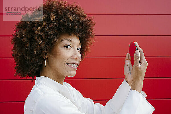 Lächelnde junge Frau hält Spiegel an roter Wand