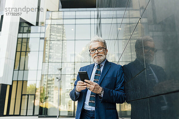 Lächelnder Geschäftsmann steht mit Smartphone an der Wand vor dem Bürogebäude