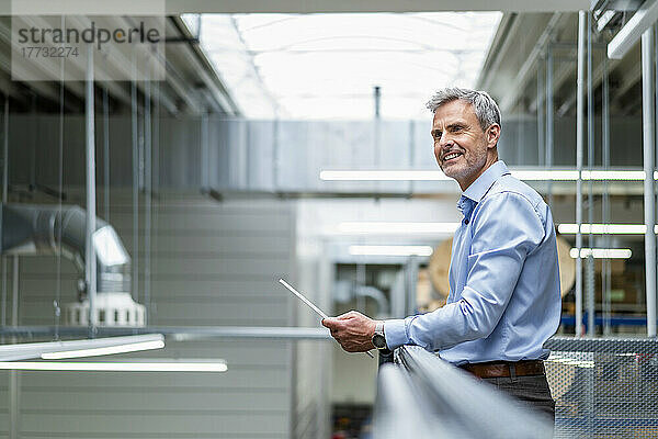 Smiling businessman leaning on railing in factory holding digital tablet