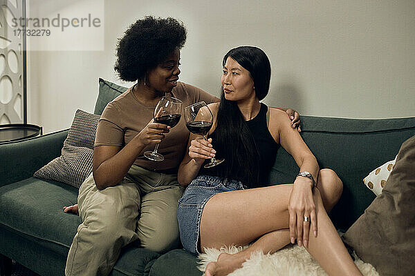 Lesbe prostet mit Weingläsern zu  während sie zu Hause auf dem Sofa im Wohnzimmer sitzt