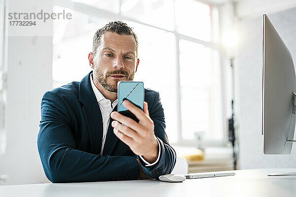 Lächelnder Geschäftsmann blickt im Büro auf sein Smartphone