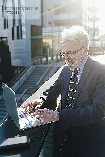 Leitender Geschäftsmann arbeitet vor dem Bürogebäude am Laptop mit Solarbatterieladegerät