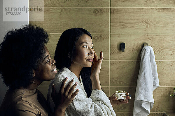 Lächelnde Frau schaut ihrer Freundin zu  die im Badezimmer Feuchtigkeitscreme auf ihr Gesicht aufträgt
