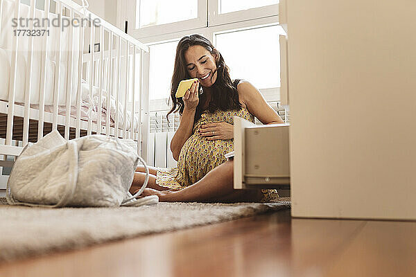 Lächelnde schwangere Frau  die zu Hause am Kinderbett sitzt und Voicemail über ihr Smartphone sendet