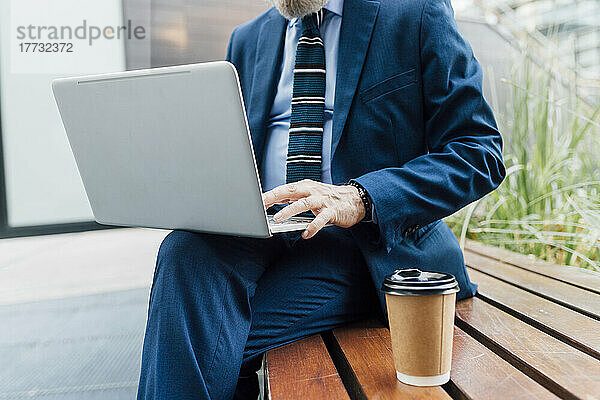 Geschäftsmann arbeitet am Laptop und trinkt eine Tasse Kaffee  während er auf einer Bank im Büropark sitzt