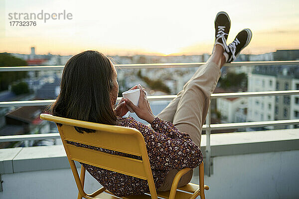 Junge Frau mit Kaffeetasse entspannt sich bei Sonnenuntergang auf dem Balkon