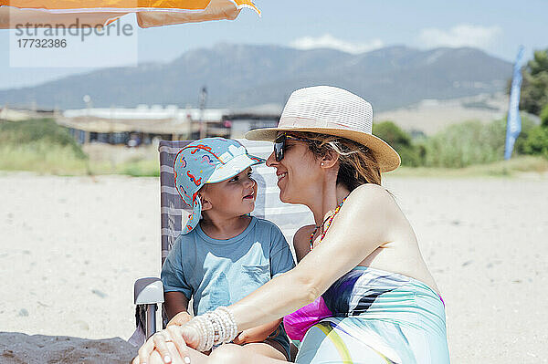 Lächelnde Frau mit Hut sitzt an einem sonnigen Tag mit ihrem Sohn am Strand