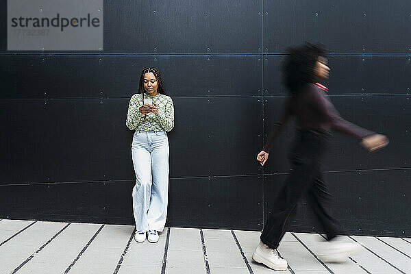 Lächelnde junge Frau  die ihr Mobiltelefon benutzt und vor der Wand steht