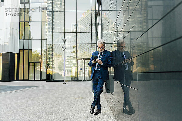 Leitender Geschäftsmann benutzt Smartphone und steht mit am Knöchel gekreuzten Beinen vor Bürogebäude