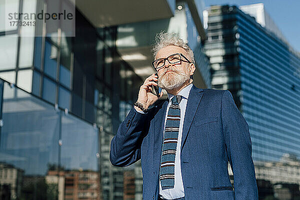 Leitender Geschäftsmann spricht mit Smartphone vor Bürogebäude