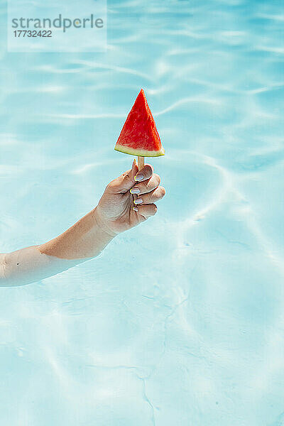 Hand einer jungen Frau hält Wassermelonenscheibe mit Eis am Stiel im Schwimmbad