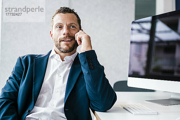 Lächelnder Geschäftsmann  der am Schreibtisch im Büro sitzt und am Smartphone spricht