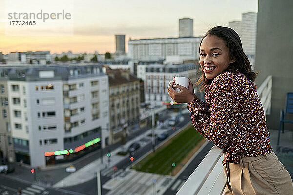 Lächelnde junge Frau mit Kaffeetasse auf dem Balkon bei Sonnenuntergang