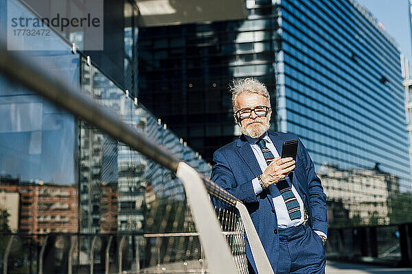 Lächelnder Geschäftsmann steht an einem sonnigen Tag mit Smartphone vor dem Bürogebäude