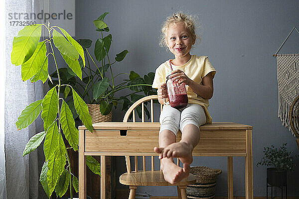 Smiling girl with jar of smoothie sitting on table at home