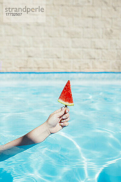 Hand einer Frau hält Wassermelonenscheibe mit Eis am Stiel im Schwimmbad