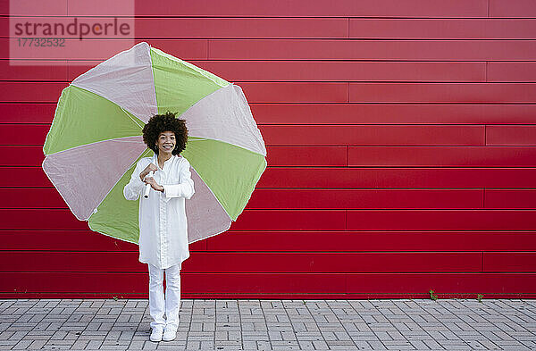 Lächelnde junge Frau mit Regenschirm steht vor roter Wand
