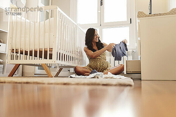Lächelnde schwangere Frau mit Babykleidung sitzt zu Hause am Kinderbett