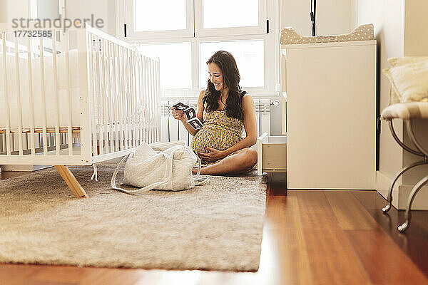Lächelnde schwangere Frau mit Ultraschalluntersuchungen  die zu Hause am Kinderbett sitzt