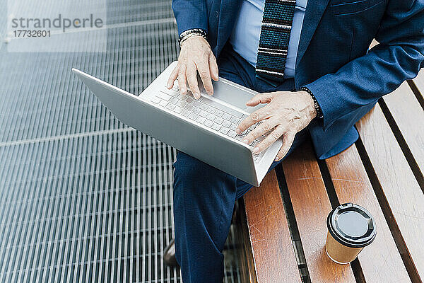 Geschäftsmann arbeitet am Laptop und sitzt auf einer Bank beim Kaffee im Freien