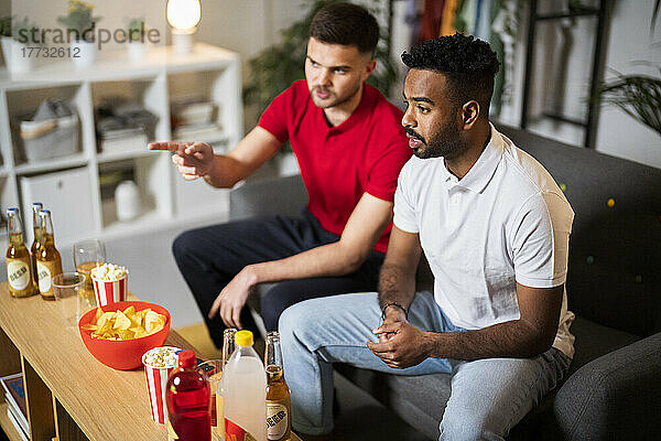 Young man talking with friend watching football match sitting on sofa at home