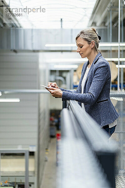 Geschäftsfrau stützt sich in Fabrik auf Geländer und nutzt digitales Tablet