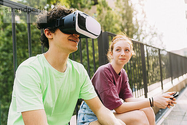 Lächelnder Junge mit Virtual-Reality-Simulator sitzt neben einem Freund vor dem Geländer