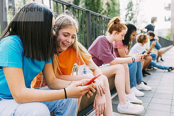 Fröhliche Mädchen  die ihr Smartphone teilen und bei Freunden vor dem Geländer sitzen