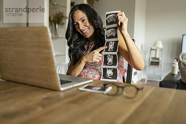 Glückliche schwangere Frau zeigt Ultraschalluntersuchungen per Videoanruf auf dem Laptop zu Hause