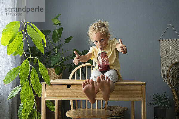 Girl drinking smoothie showing thumbs up gesture sitting on table