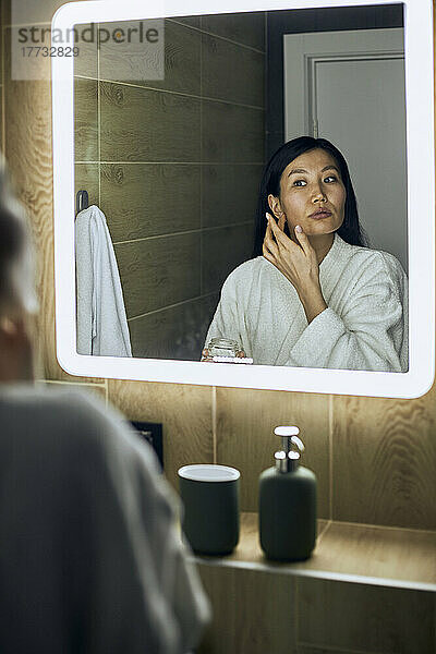 Frau im Bademantel trägt Feuchtigkeitscreme auf ihr Gesicht auf und blickt in den Spiegel