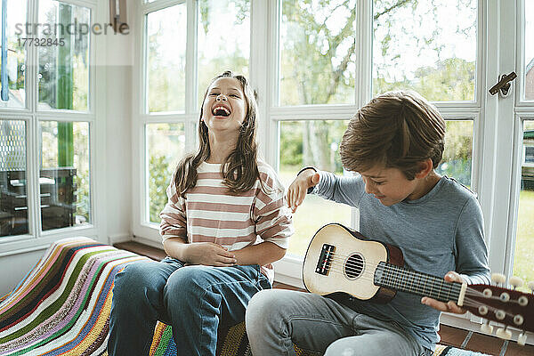Fröhliches Mädchen sitzt mit Bruder und spielt zu Hause Gitarre