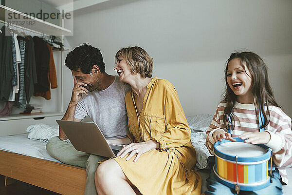 Glückliche Männer und Frauen sitzen mit Laptop neben ihrer Tochter und spielen zu Hause im Bett Spielzeugtrommel