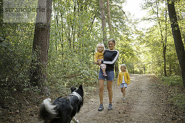Mutter trägt Tochter und geht mit Mädchen und Hund im Wald spazieren