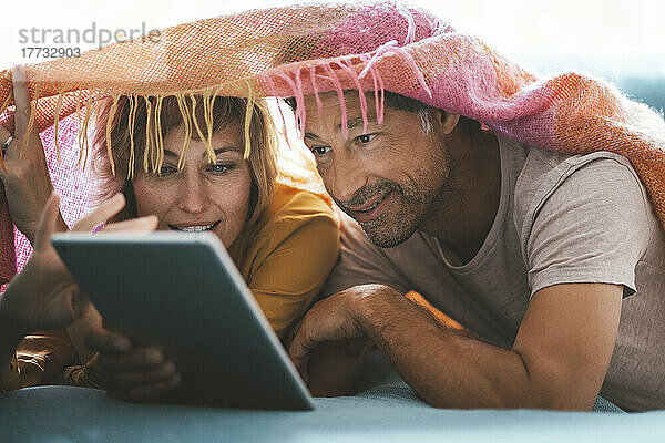 Reifer Mann und Frau mit gemeinsamem Tablet-PC liegen zu Hause unter der Decke