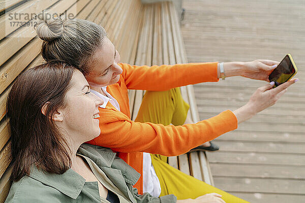Lächelnde Frau macht ein Selfie mit einer Freundin per Smartphone  die auf einer Bank sitzt