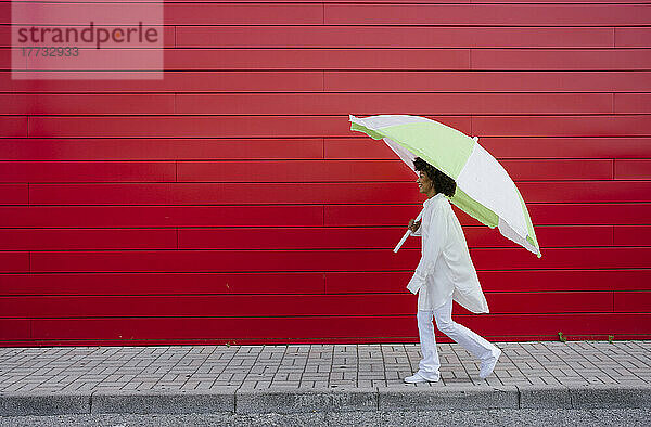 Young woman walking with umbrella on footpath by red wall