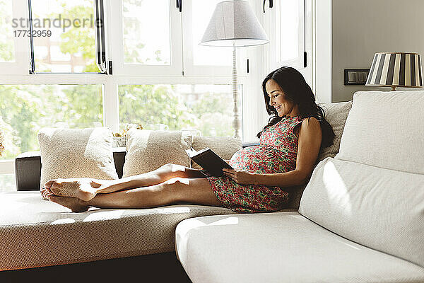 Lächelnde schwangere Frau  die zu Hause auf dem Sofa im Wohnzimmer ein Buch liest