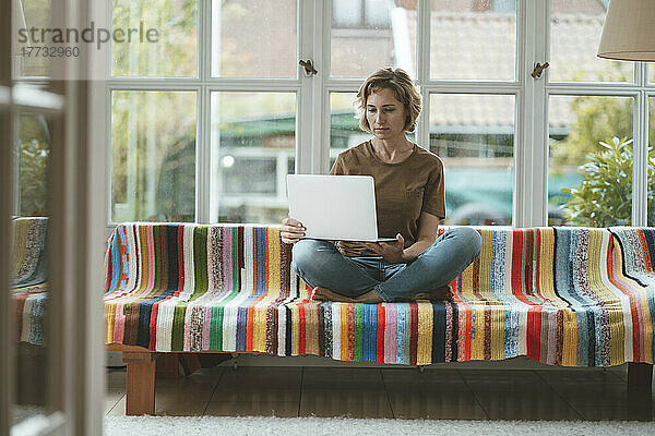 Frau sitzt mit Laptop auf Sofa vor Fenster