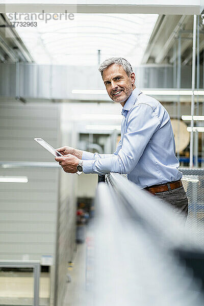 Lächelnder Geschäftsmann lehnt an Geländer in Fabrik und hält digitales Tablet in der Hand