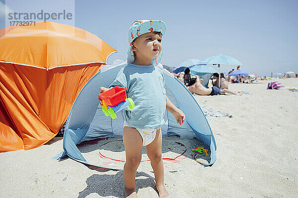 Junge hält Spielzeugauto und steht an einem sonnigen Tag vor Zelt am Strand