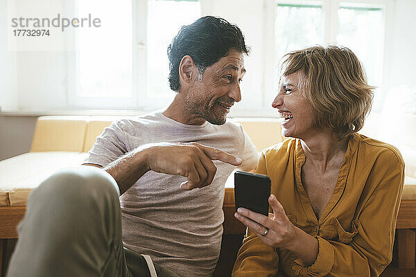 Lächelnder Mann unterhält sich mit einer fröhlichen Frau  die zu Hause ihr Smartphone hält