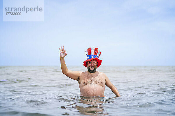 Lächelnder  hemdloser  fettleibiger Mann mit Uncle-Sam-Hut  der im Meer ein OK-Schild zeigt