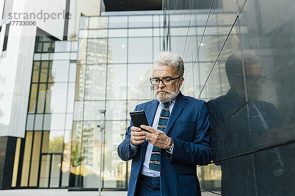 Leitender Geschäftsmann arbeitet vor dem Bürogebäude am Smartphone