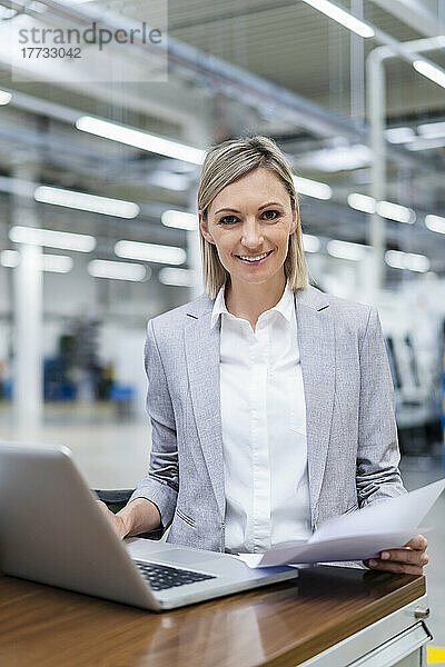 Porträt einer lächelnden Geschäftsfrau mit Laptop und Dokumenten in der Fabrik