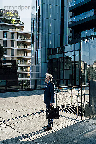 Leitender Geschäftsmann steht mit Aktentasche vor der Fassade des Bürogebäudes