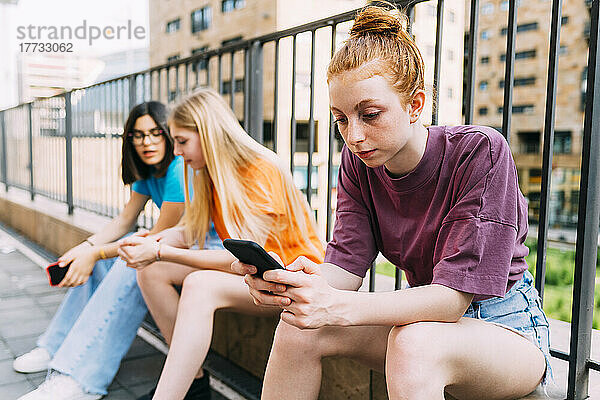 Mädchen mit blonden Haaren sitzt mit Freunden vor dem Geländer und benutzt Smartphone