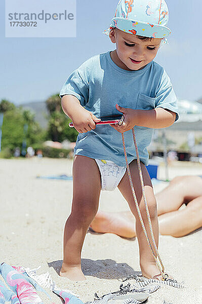Netter Junge mit Smartphone steht auf Sand am Strand