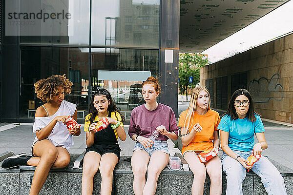 Multiracial friends eating snacks sitting in front of building