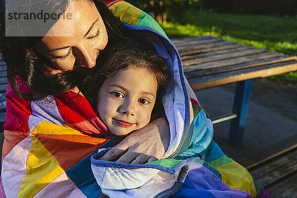 Mutter und Tochter sitzen auf einer Bank  in eine Steppdecke gehüllt  im Park